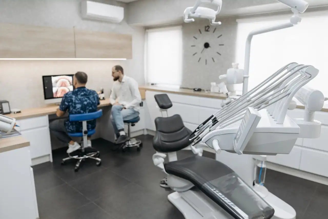 Cabinet dentaire moderne avec fauteuil dentaire en premier plan, deux praticiens consultant un écran de radiographie, mobilier blanc et bois, éclairage professionnel